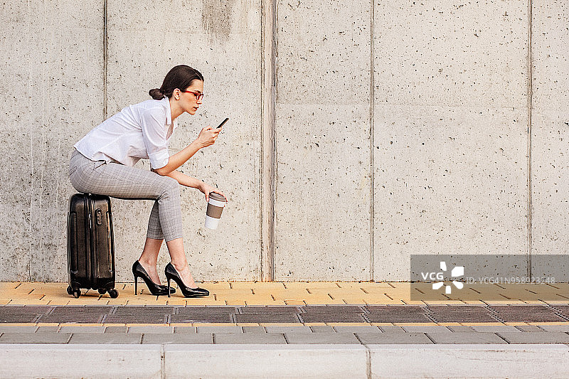 坐在人行道行李箱上使用智能手机的年轻女子图片素材