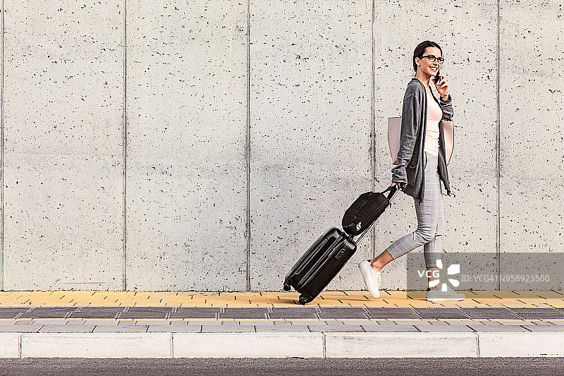 年轻女人走在水泥墙边，拉着带公文包的小型轮式行李箱，同时使用智能手机图片素材