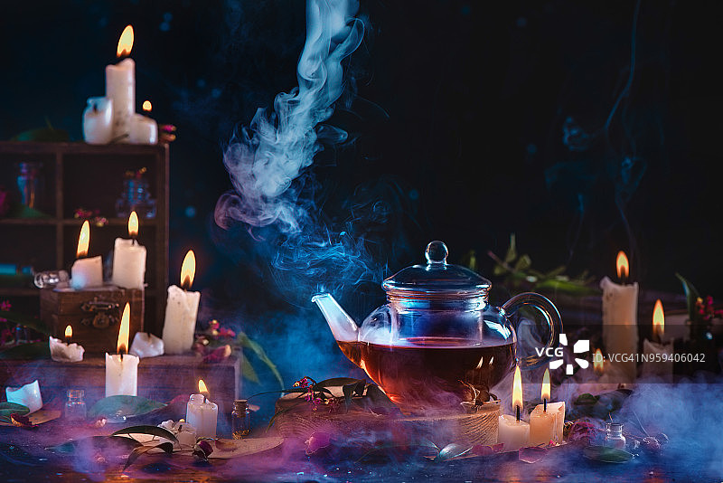 深色静物中带有上升蒸汽的玻璃茶壶，燃烧的蜡烛和烟雾。制作草药茶概念，带有复制空间。图片素材