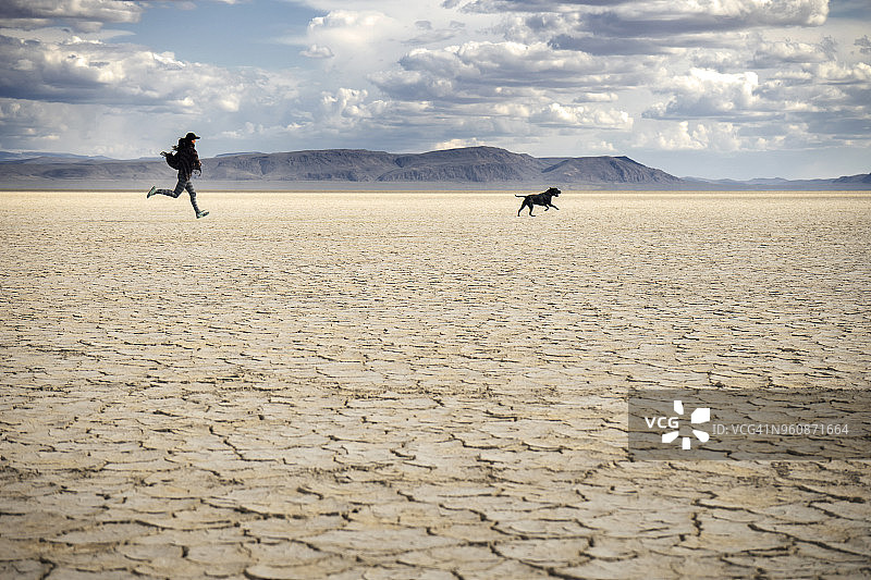 在阿尔沃德沙漠，女人和狗在干燥的田野上奔跑，天空多云图片素材