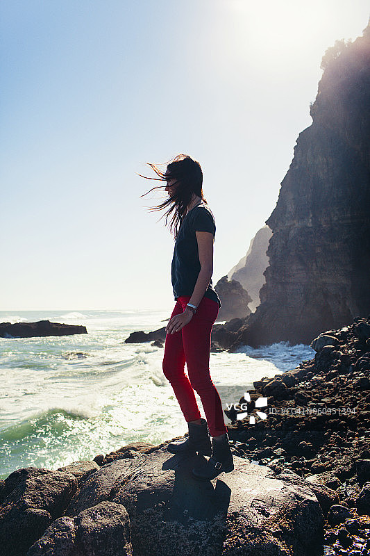 女子在晴朗的日子里站在岸边的岩石上，面朝大海和天空图片素材