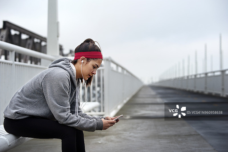 坐在海湾大桥上用智能手机听音乐的女性运动员侧视图图片素材