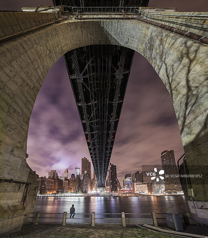 夜晚，男子坐在皇后区大桥下的栏杆上，以多云的天空为背景的背影图片素材