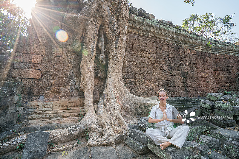 年轻女人在古庙里靠近雄伟的树根处锻炼瑜伽；阳光穿过，通过冥想放松身心；健康生活方式、幸福和旅行的概念图片素材
