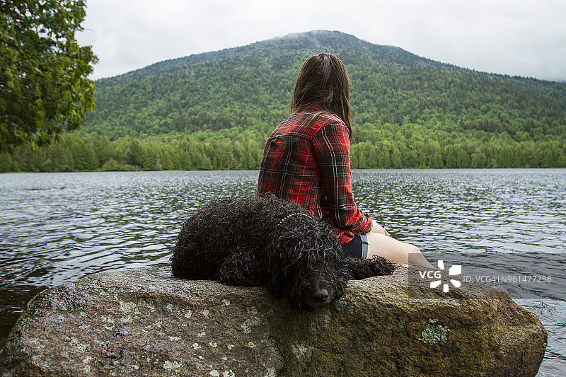 女子坐在森林岩石上，小狗在旁边睡觉，女子看着池塘的侧视图图片素材