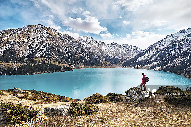 背包客在冬季站在湖边欣赏山景和多云的天空图片素材