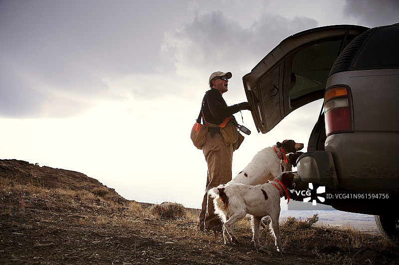 猎人与猎犬站在汽车旁的侧视图，背景是天空图片素材
