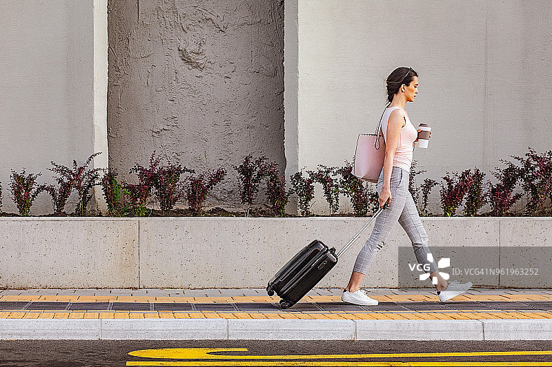 年轻女子走在混凝土墙旁的人行道上，拉着一个带轮子的小行李箱图片素材