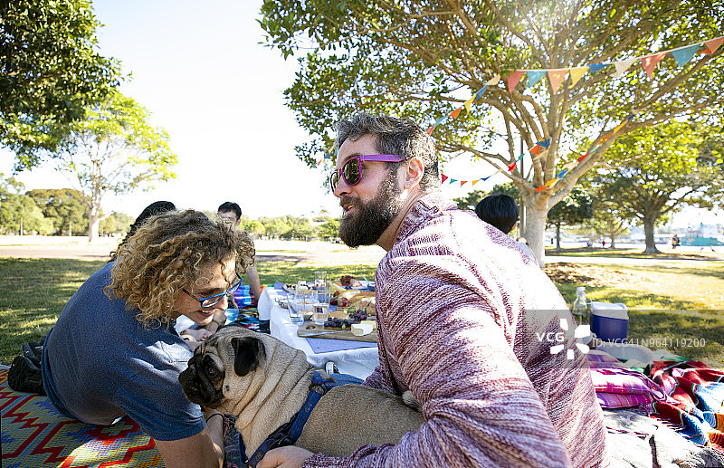 在公园的野餐垫上与巴哥犬玩耍的两个男人图片素材