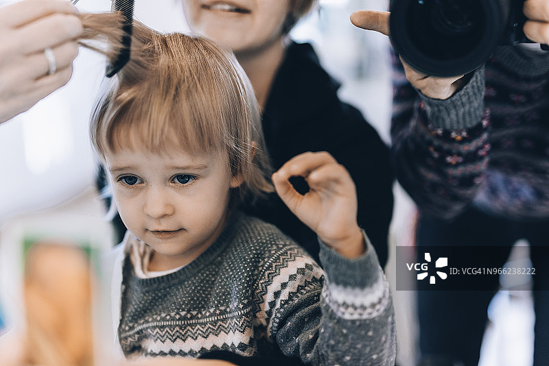 男人在沙龙里拍摄理发师给坐在母亲身边的女孩理发图片素材