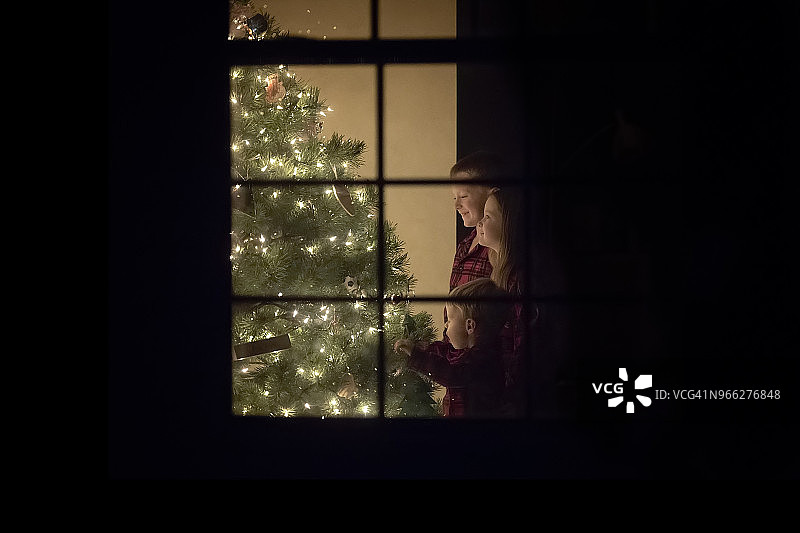 微笑的兄弟姐妹站在家中被照亮的圣诞树旁，透过窗户看到图片素材
