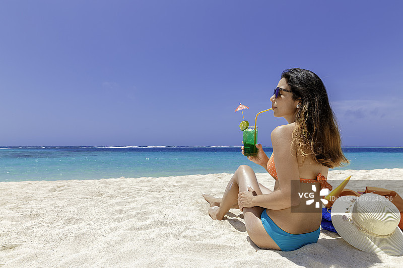 西班牙裔年轻女子在加勒比海热带白色沙滩上享受日光浴，并品尝鸡尾酒图片素材