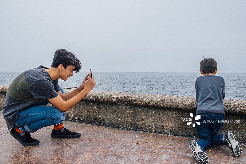 青少年男孩用带摄像头的手机拍摄木板路上户外玩耍的幼童图片素材