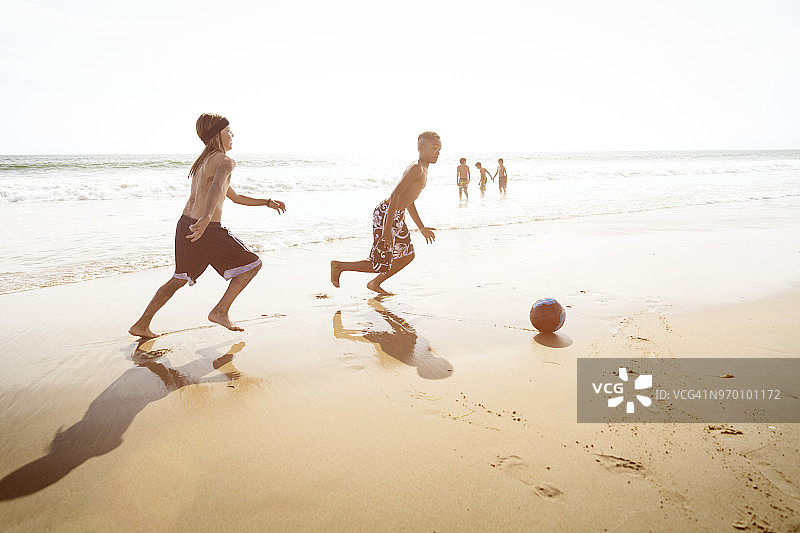 男孩们在海滩上踢足球的侧面视角，背景是晴朗的天空图片素材
