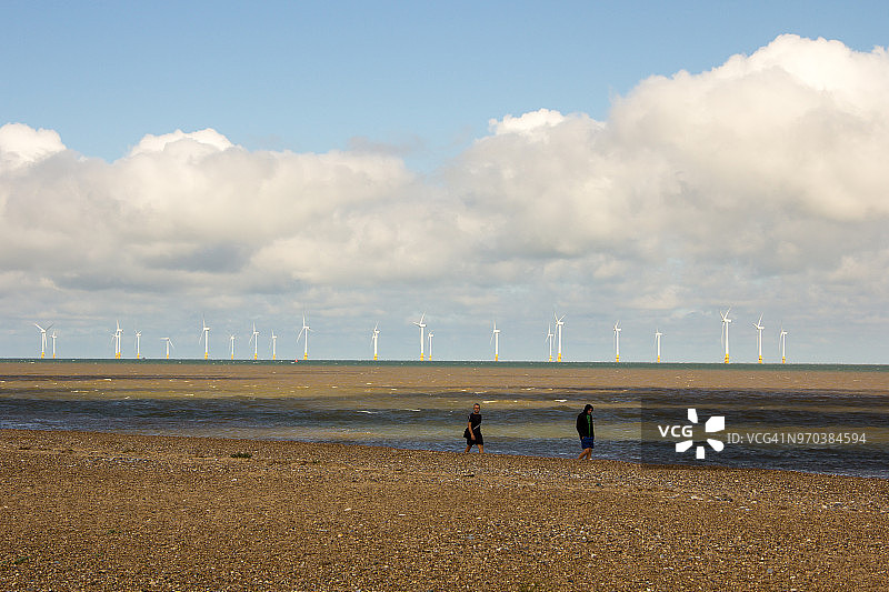 两人走在英国诺福克郡大雅茅斯卵石滩上，背景是距岸2.5公里的斯克罗比沙洲风电场图片素材