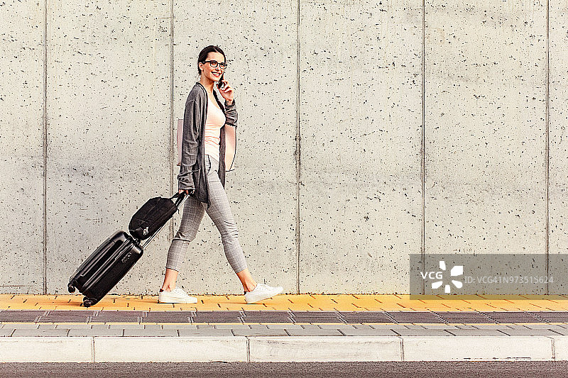 年轻女人走在水泥墙边，拉着带公文包的小型轮式行李箱，同时使用智能手机图片素材