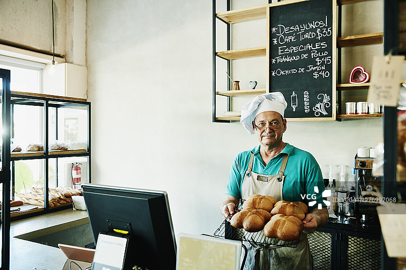 面包师站在商店柜台后，手持一盘新鲜出炉的面包图片素材