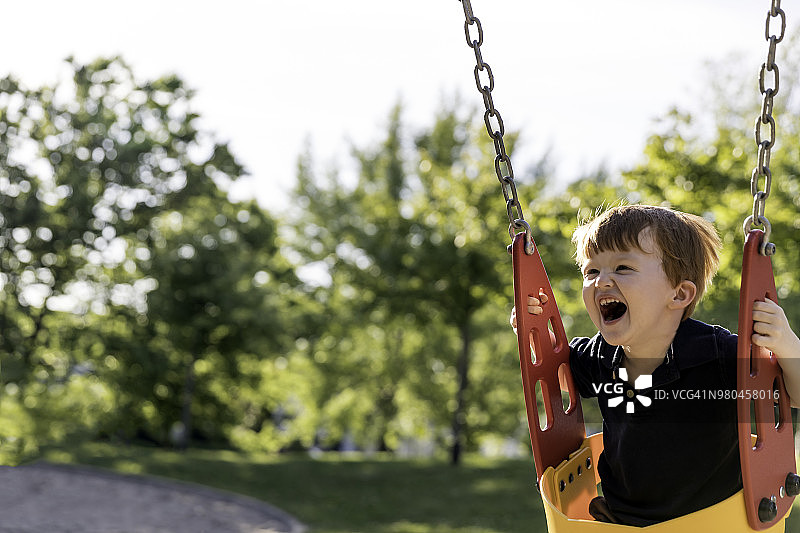 在户外游乐场秋千上快乐的红发幼儿男孩图片素材