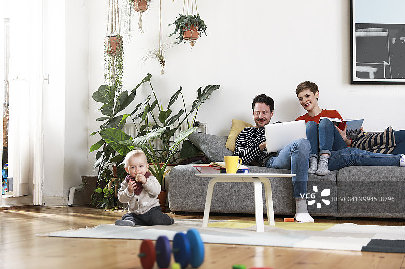 父母坐在沙发上使用笔记本电脑，女儿在地板上玩耍图片素材