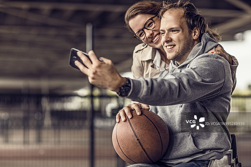 使用轮椅的年轻男子在户外城市篮球训练期间与女友自拍图片素材