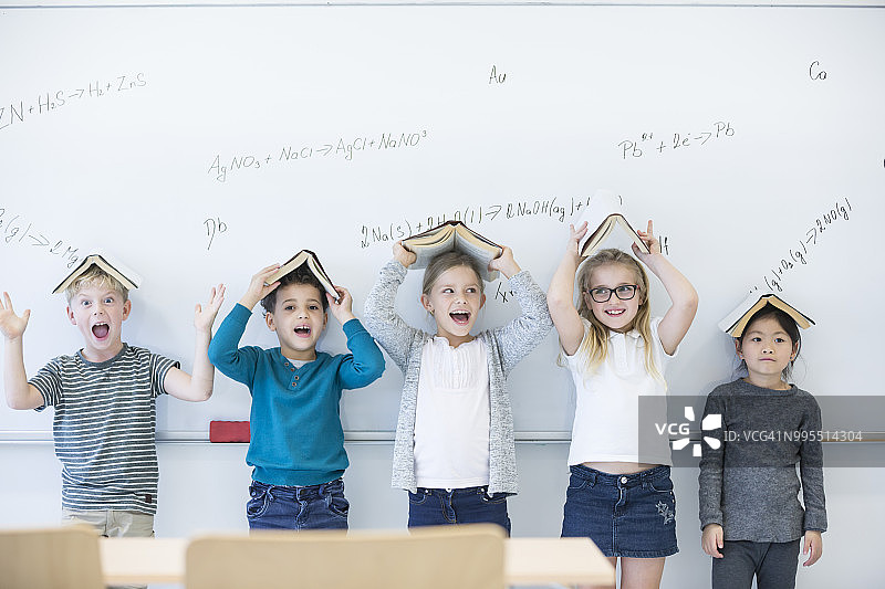 课堂上，快乐的小学生们头顶书本站在写满公式的白板前图片素材