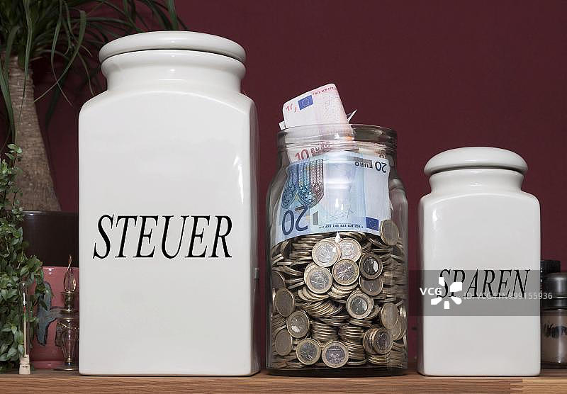 厨房架子上装有钱的罐子，以及标有“税”和“储蓄”字样的罐子，德国图片素材