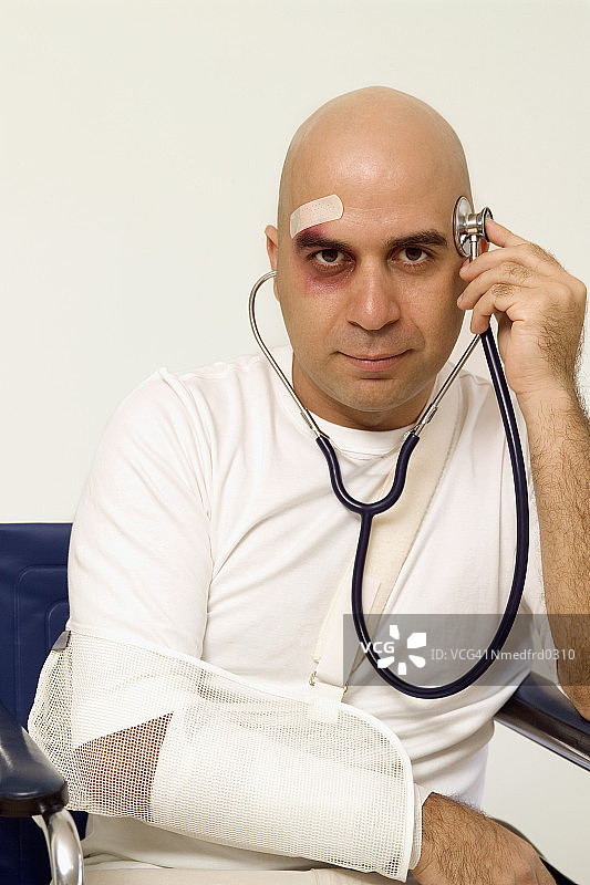 特写：额头上有绷带、手臂吊着绷带、眼睛发黑，拿着听诊器的光头男人图片素材