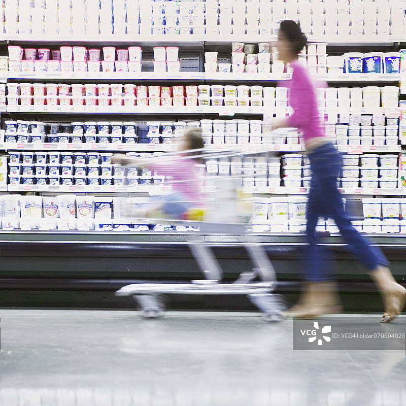 妈妈在超市通道里推着坐在购物车里的女儿（模糊）图片素材