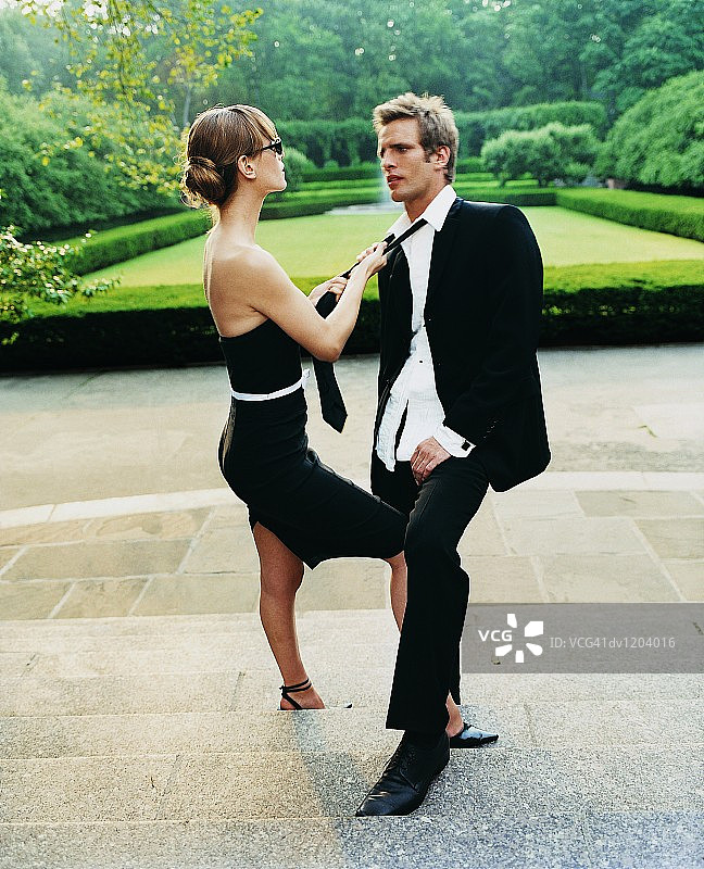 戴墨镜穿晚礼服的年轻女子在花园台阶上拉着男人的领带图片素材