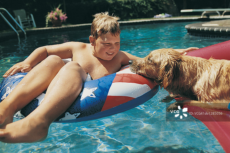 男孩坐在游泳池的充气床上，旁边有他的狗图片素材