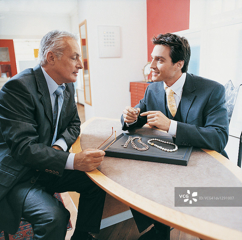 男人坐在珠宝店的桌子旁，对面是店员，看着项链和戒指图片素材