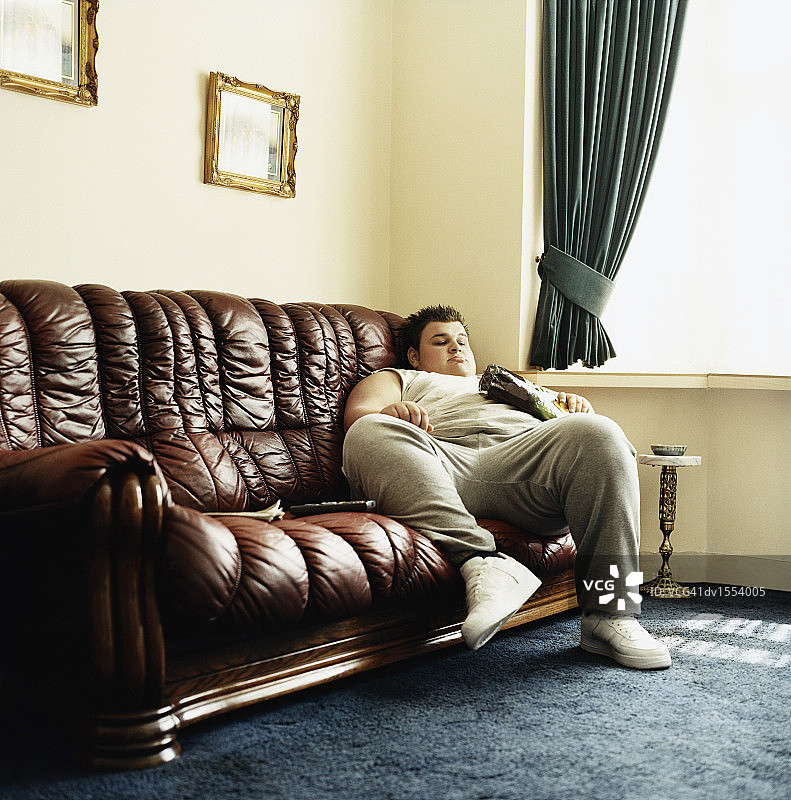 年轻男人躺在沙发上看电视，手里拿着一包薯片图片素材