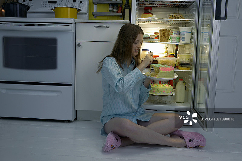 穿着睡衣坐在厨房地板上，在冰箱旁边吃蛋糕的女人图片素材