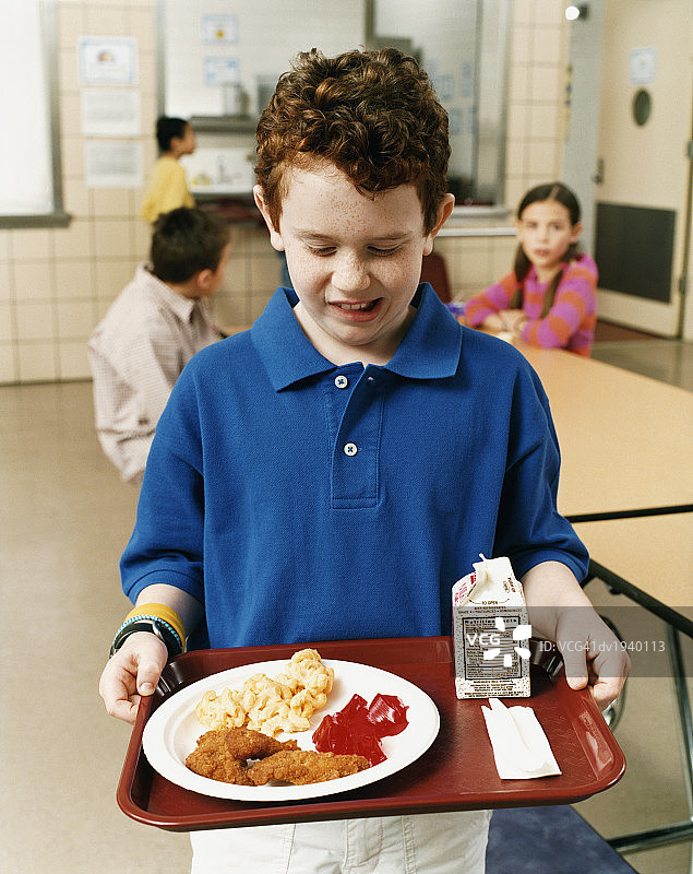 小学生站在学校食堂里，拿着不健康的学校午餐图片素材