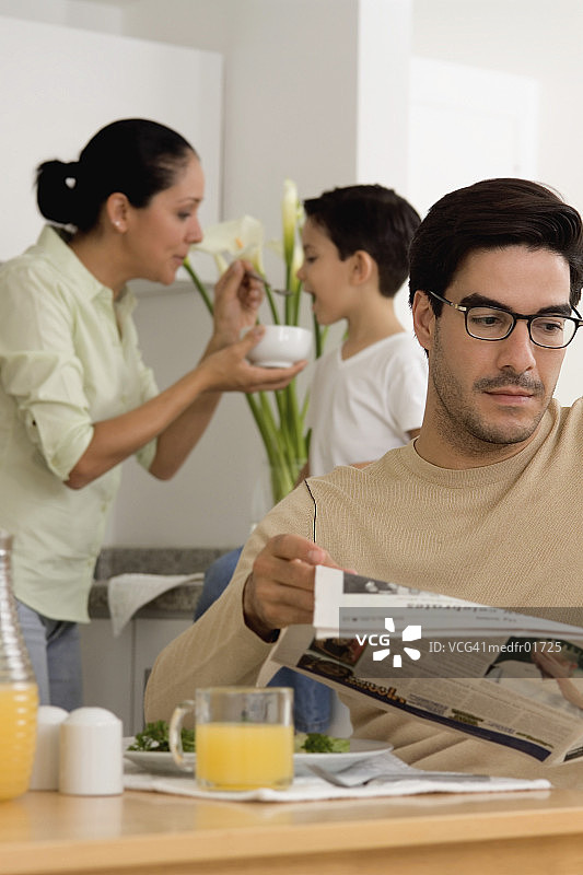 男人坐在桌旁看报纸，女人在厨房喂男孩图片素材