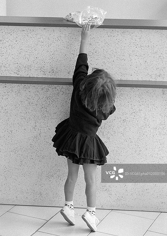 黑白照片：踮起脚尖，伸手去拿架子上塑料袋的小女孩图片素材