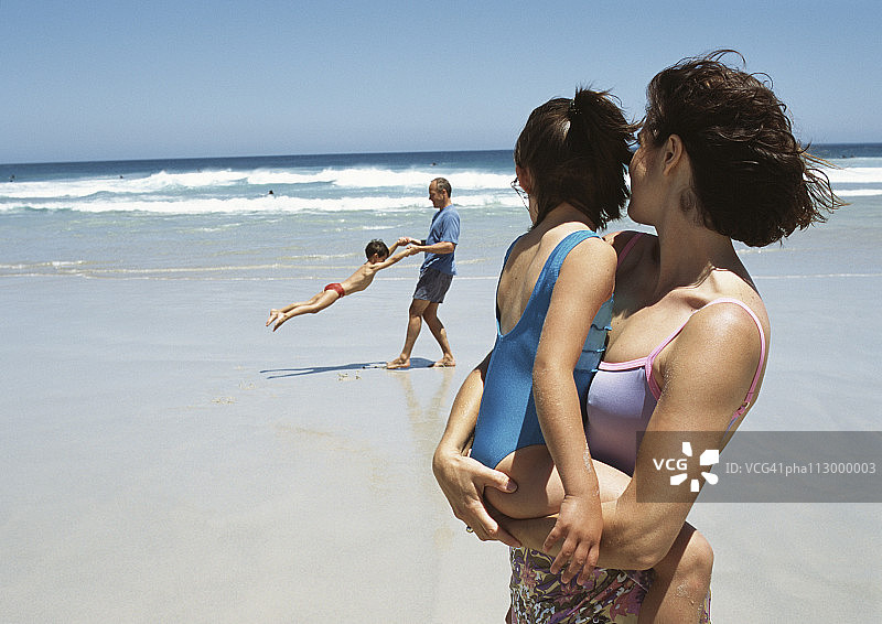 海滩上，女人怀抱小女孩，男人在背景中摇晃小男孩图片素材