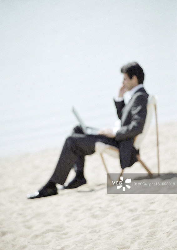 坐在沙滩上，膝盖上放着笔记本电脑的商人（全身特写，模糊）图片素材