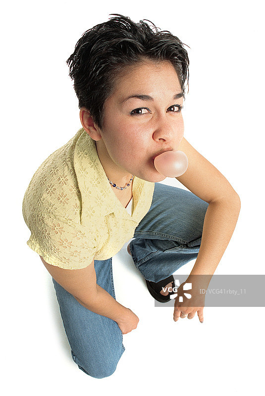 年轻白人女孩梳着深色头发，穿着蓝色牛仔裤和黄色上衣，吹着泡泡糖图片素材