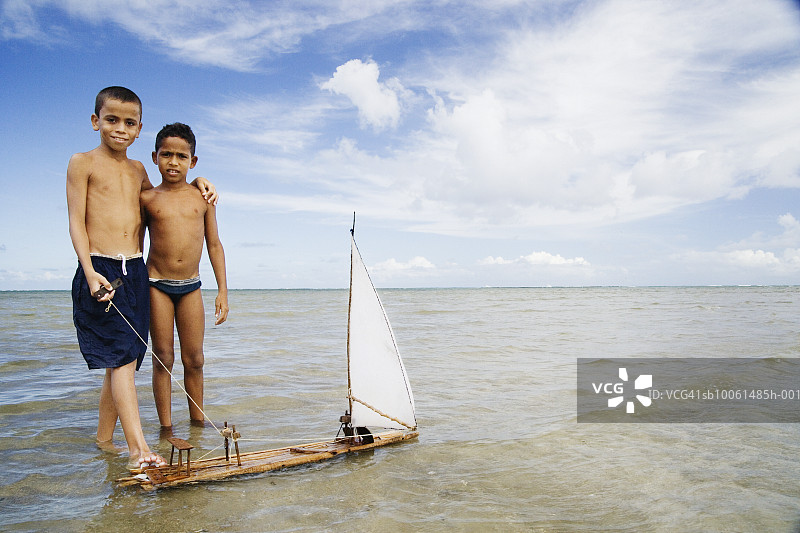 两个年轻的男孩（8-9岁，10-11岁）站在海边浅水中，拿着手工制作的船图片素材