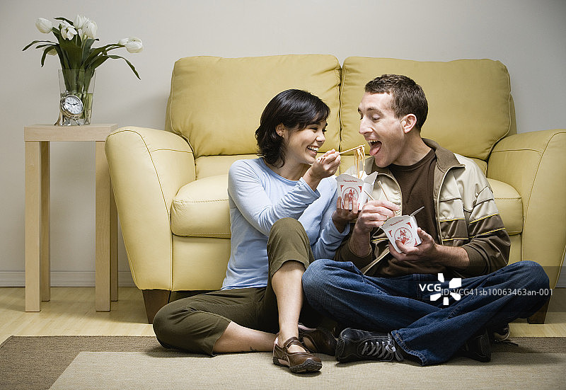 年轻夫妇盘腿坐在沙发前吃外卖，女人用筷子喂男人图片素材