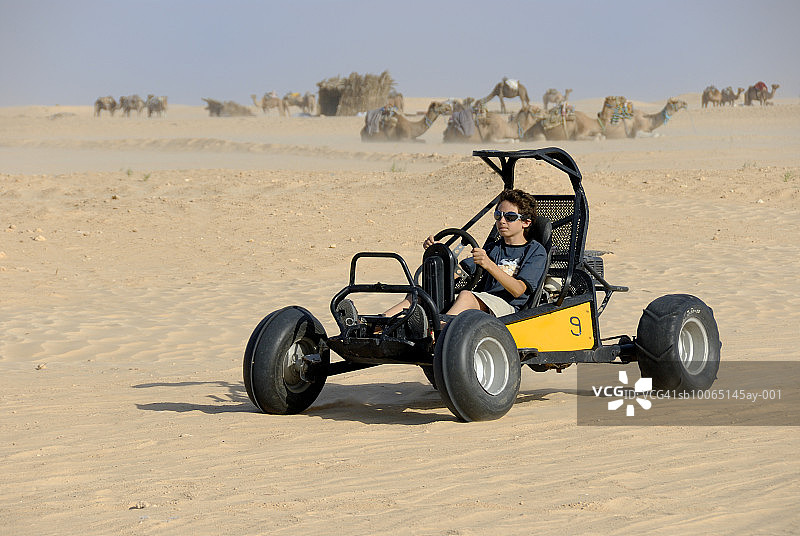 男孩（12-13岁）在沙漠中驾驶沙滩车，背景是骆驼图片素材