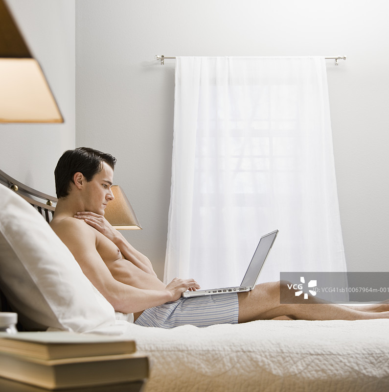 男人坐在床上使用笔记本电脑，侧视图图片素材