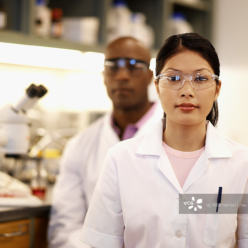 年轻女微生物学家佩戴防护眼镜的肖像，一位年轻男微生物学家坐在她身后图片素材