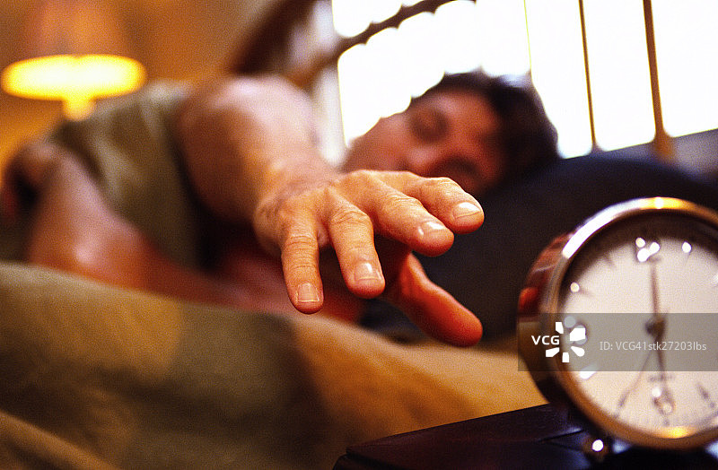 低角度拍摄：一个年轻人躺在床上，伸手去拿闹钟图片素材