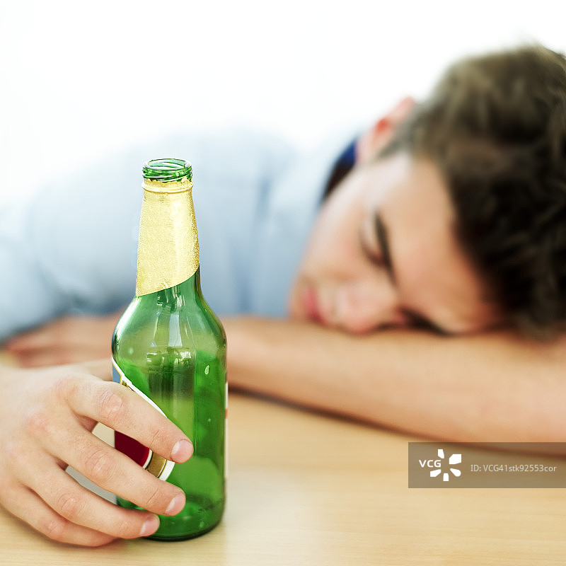 年轻男人趴在桌子上睡着了，手里拿着一个空啤酒瓶图片素材