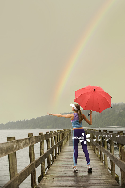 穿着运动服、在码头上撑着红色雨伞追逐彩虹的白人女性图片素材